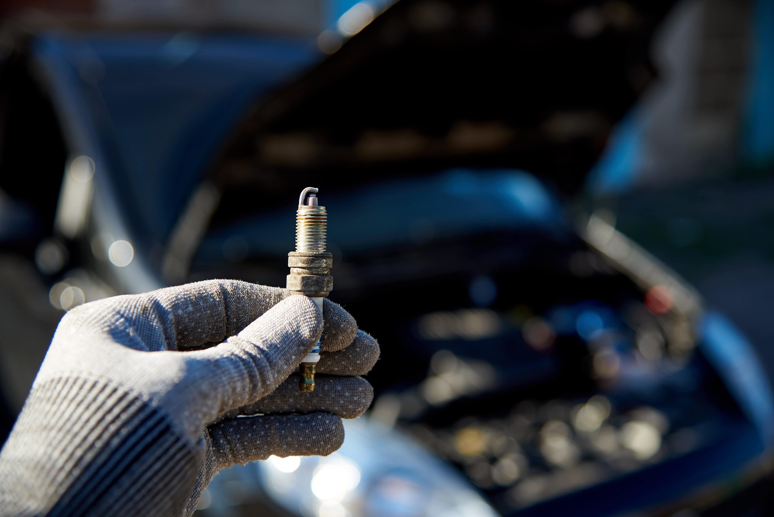 Person holding a sparkplug with a glove in front of an open engine hood