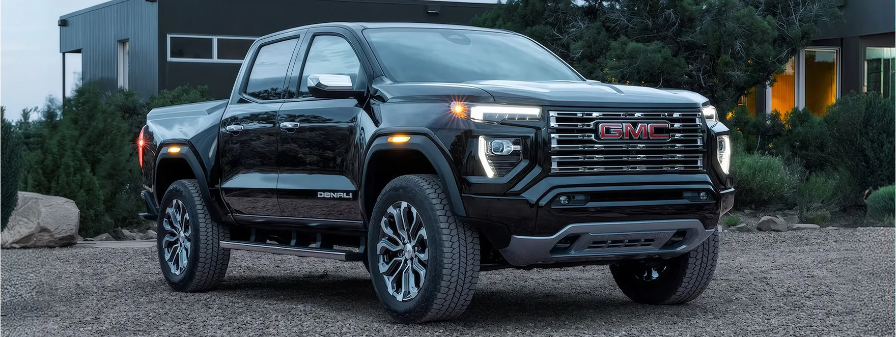 Black GMC Canyon Denali parked on gravel beside a modern building, seen from the front three-quarter angle