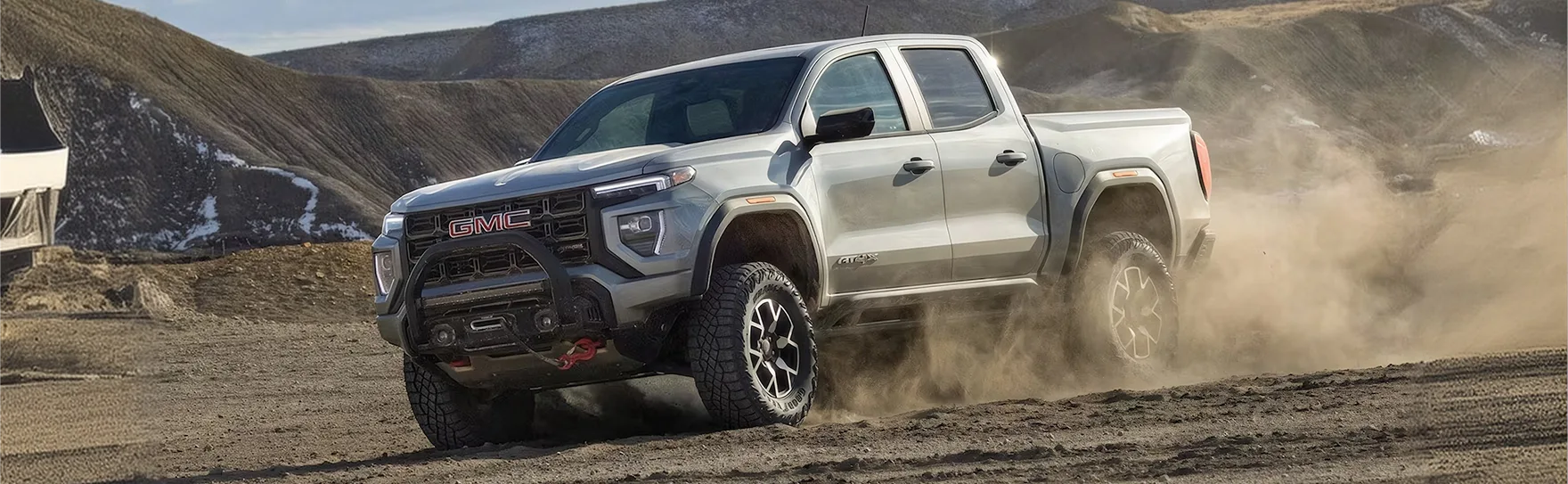 White GMC Canyon AT4X driving hard across a dirt trail, throwing up a cloud of dust
