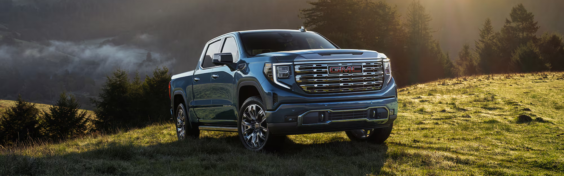 Blue GMC Sierra 1500 Denali on a grassy hillside with mist in the valley behind it