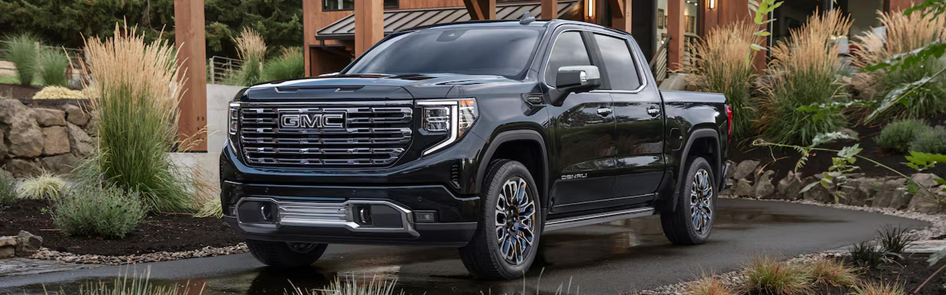 Black GMC Sierra 1500 Denali parked on a driveway beside a timber-framed home, seen from the front three-quarter angle