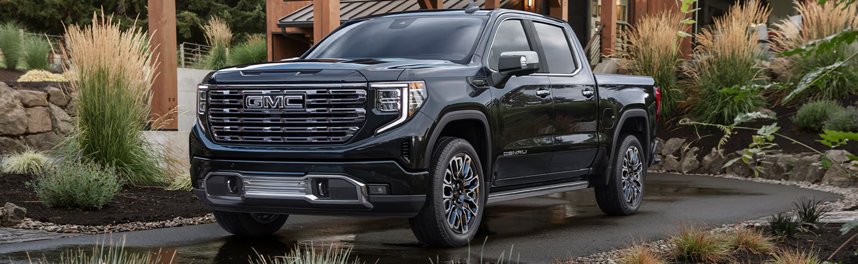 Black GMC Sierra 1500 Denali parked on a wet driveway among ornamental grasses, seen from the front three-quarter angle