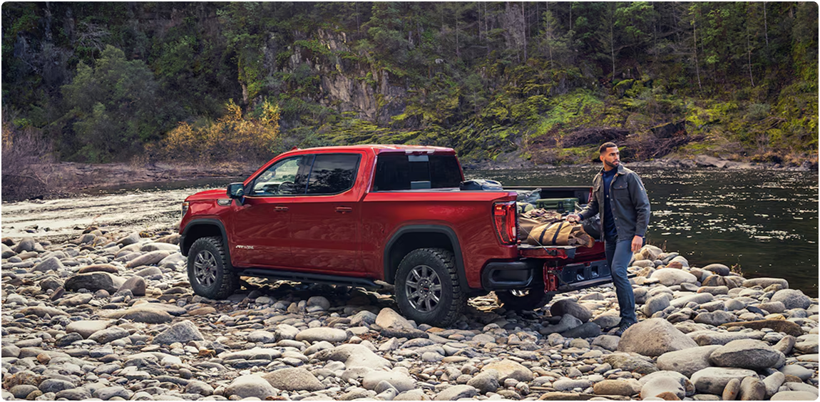 Red GMC Sierra 1500 AT4X parked on a rocky riverbank with a man standing at the open tailgate
