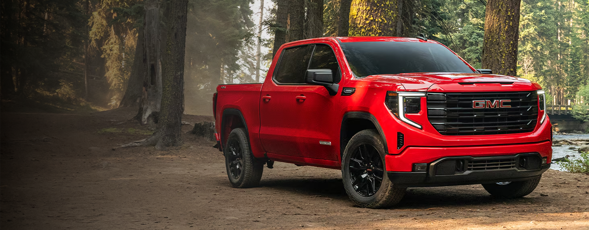 Red 2026 GMC Sierra 1500 Elevation parked on a forest dirt road beside a stream, seen from the front three-quarter angle