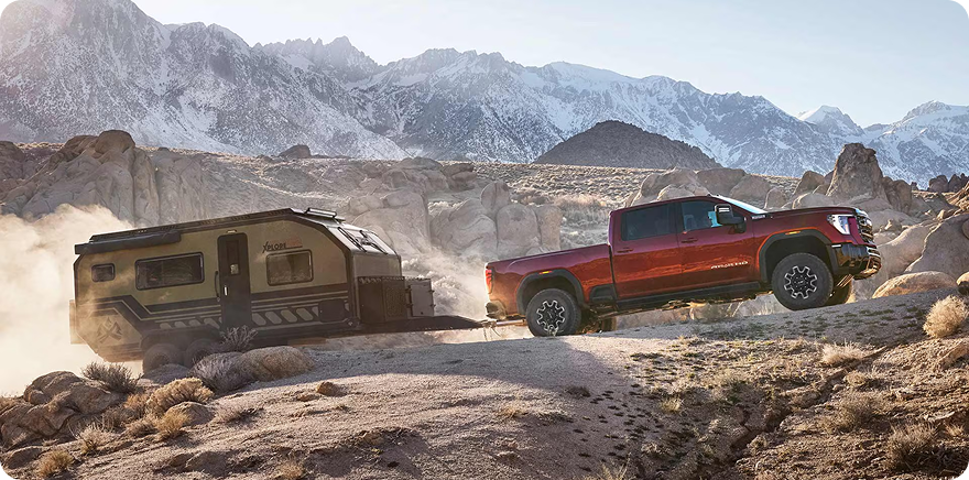 Red GMC Sierra HD towing a camping trailer up a rocky desert trail with snow-capped mountains behind
