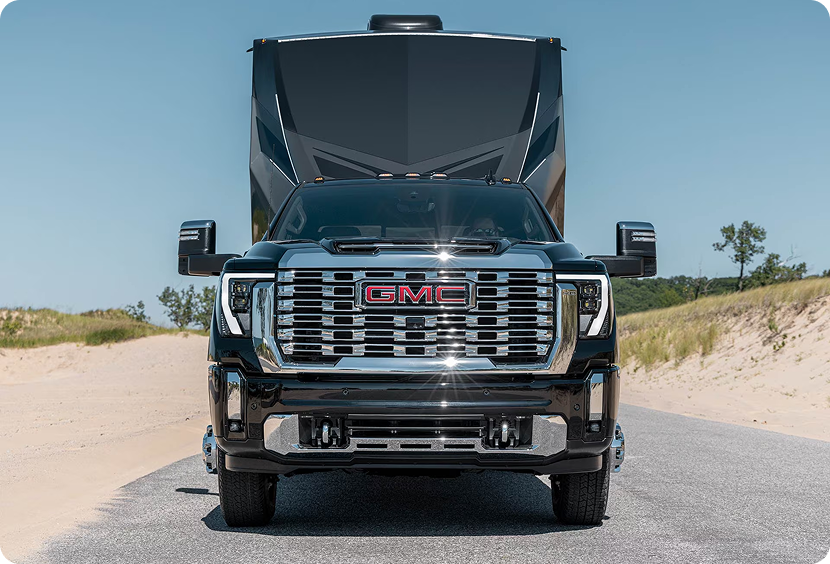 Front view of a black GMC Sierra HD Denali with a chrome grille towing a fifth-wheel trailer on a sandy road