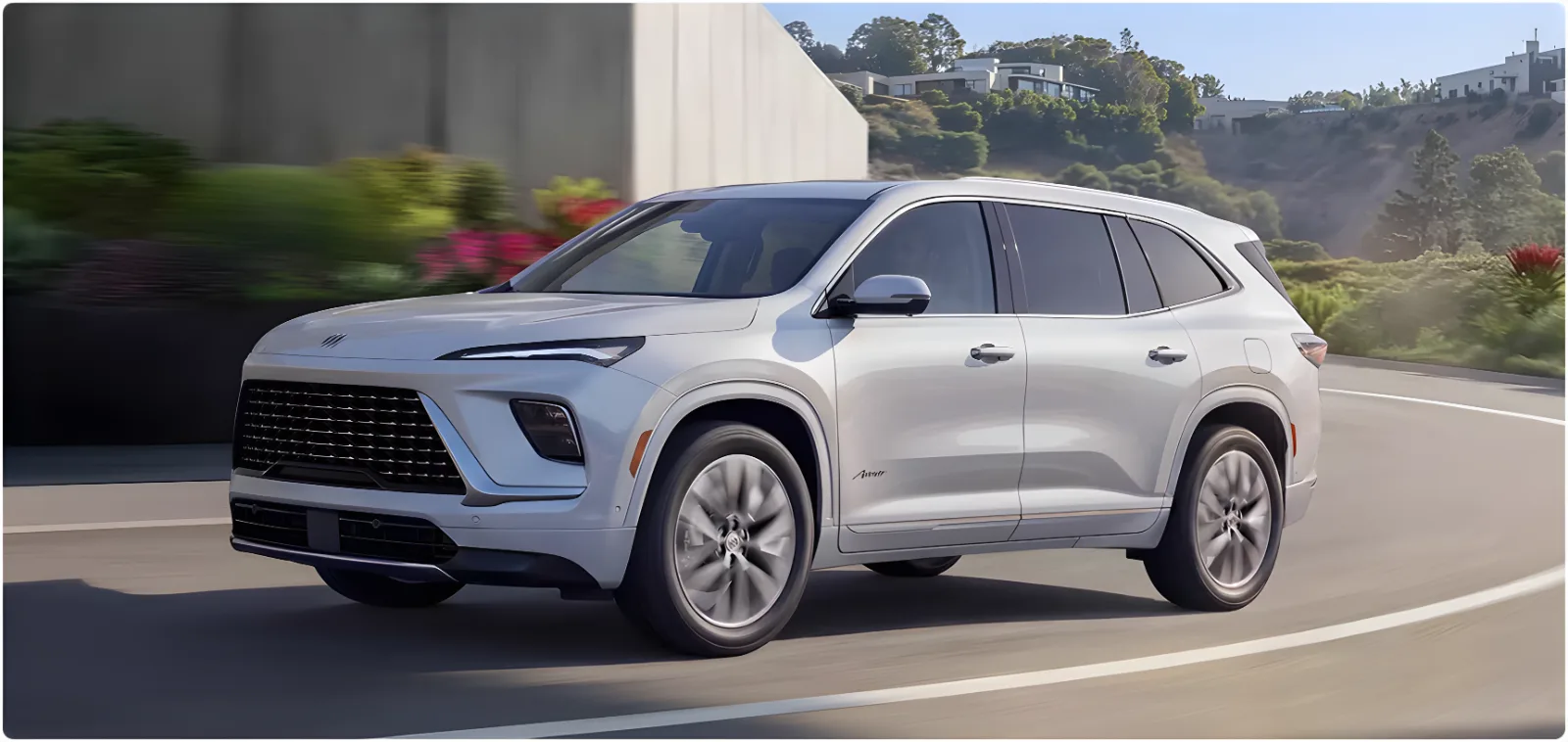 White Buick Enclave driving a tree-lined road, seen from the front three-quarter angle