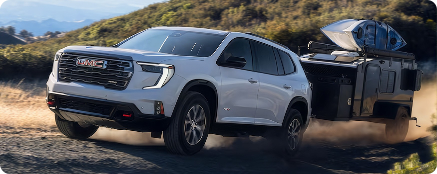 White GMC Acadia AT4 towing a camping trailer with kayaks along a dusty road
