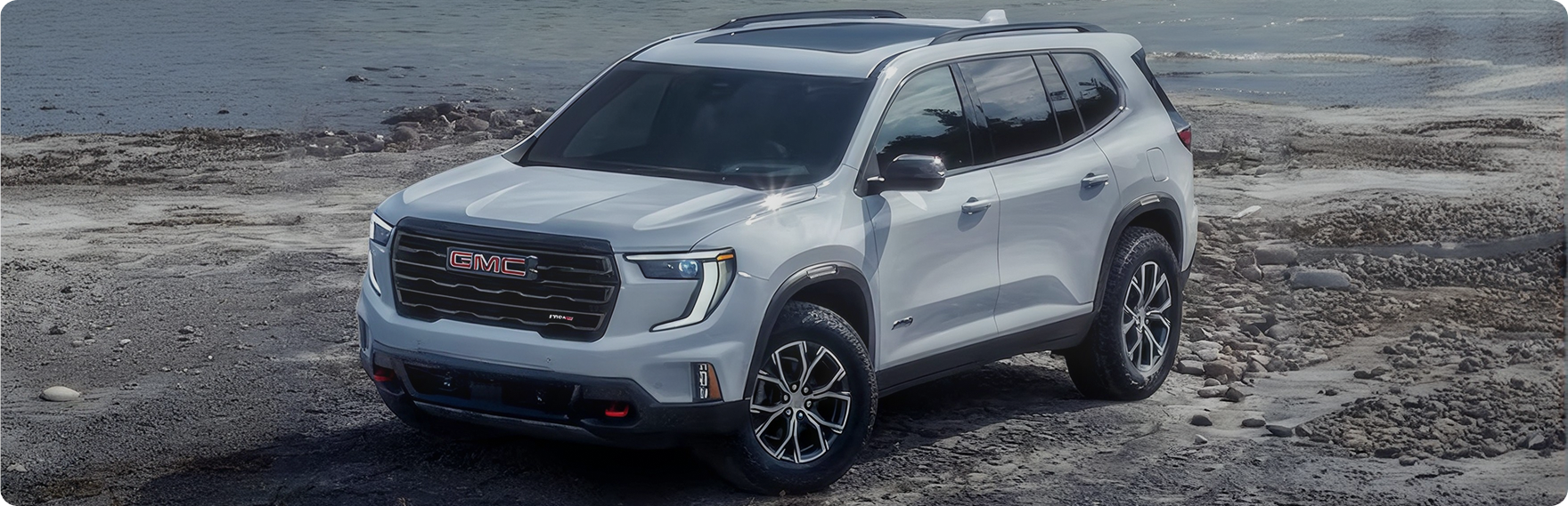 White GMC Acadia AT4 parked on a rocky shoreline, seen from the front three-quarter angle
