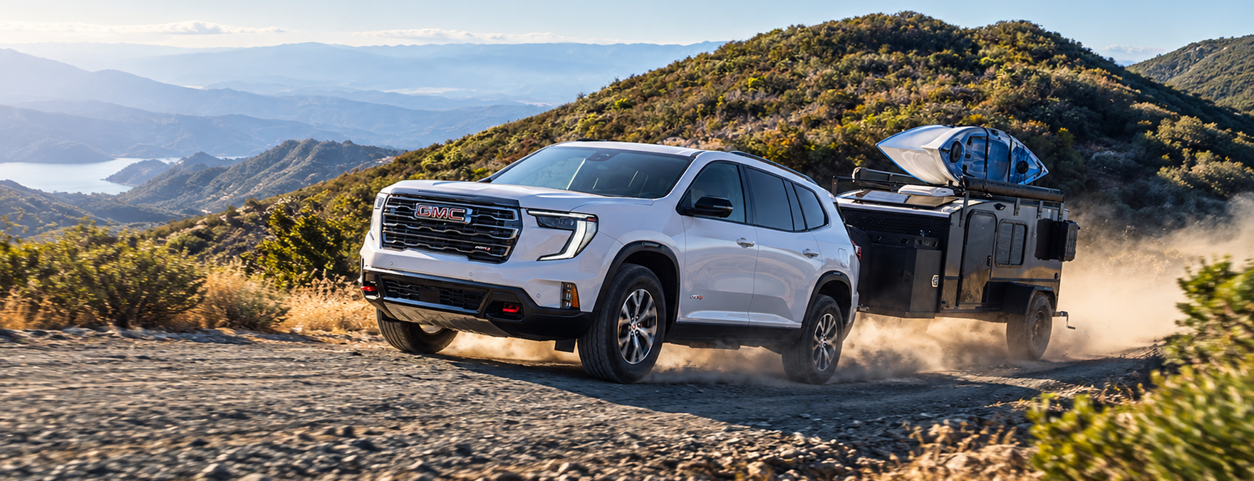 White GMC Acadia AT4 towing a camping trailer on a mountain road above a lake