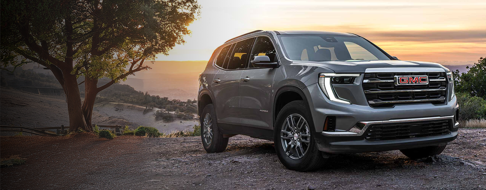 Gray 2027 GMC Acadia parked on a dirt overlook at sunset, seen from the front three-quarter angle