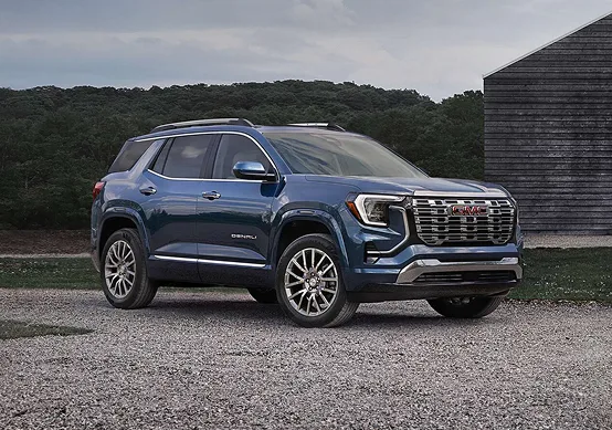 Blue 2027 GMC Terrain Denali beside a dark barn, showing its Galvano chrome grille and chrome side accents