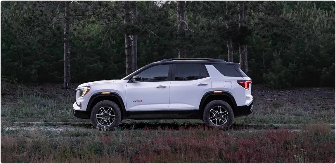 White 2027 GMC Terrain parked in profile among tall pines at dusk