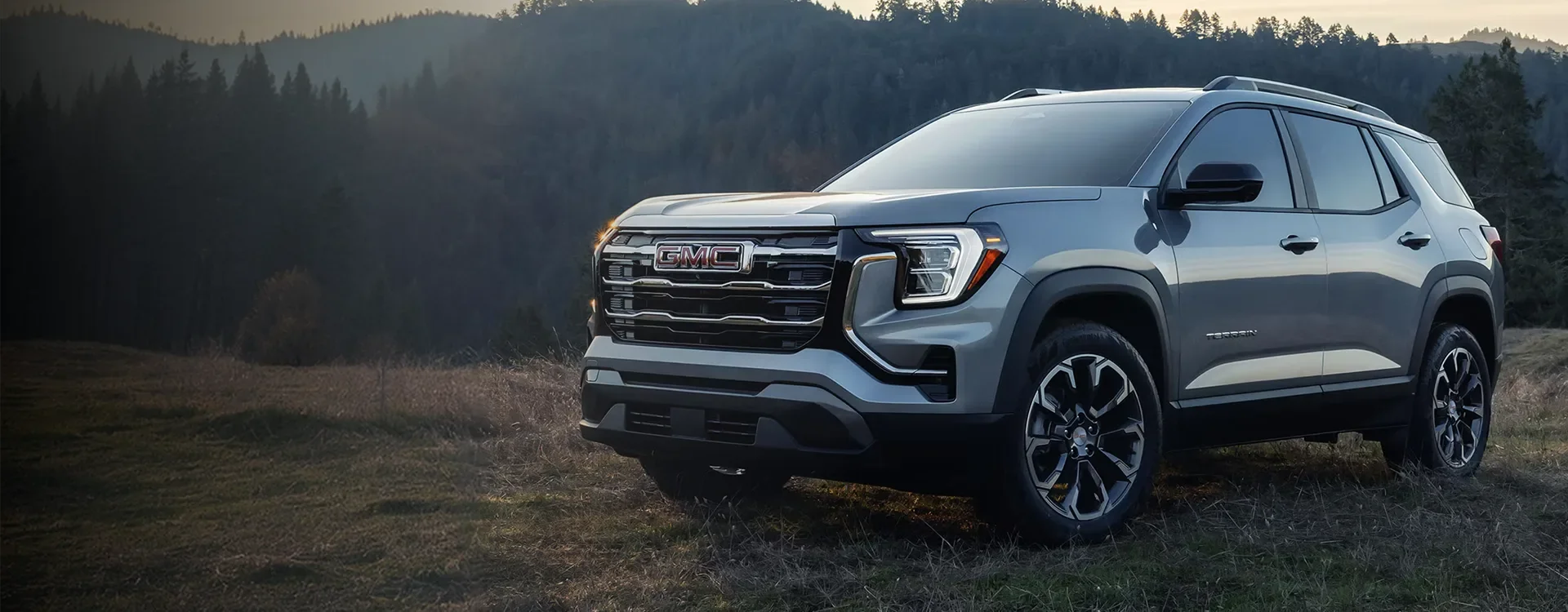 Grey 2027 GMC Terrain parked in a mountain meadow at sunrise, seen from the front three-quarter angle