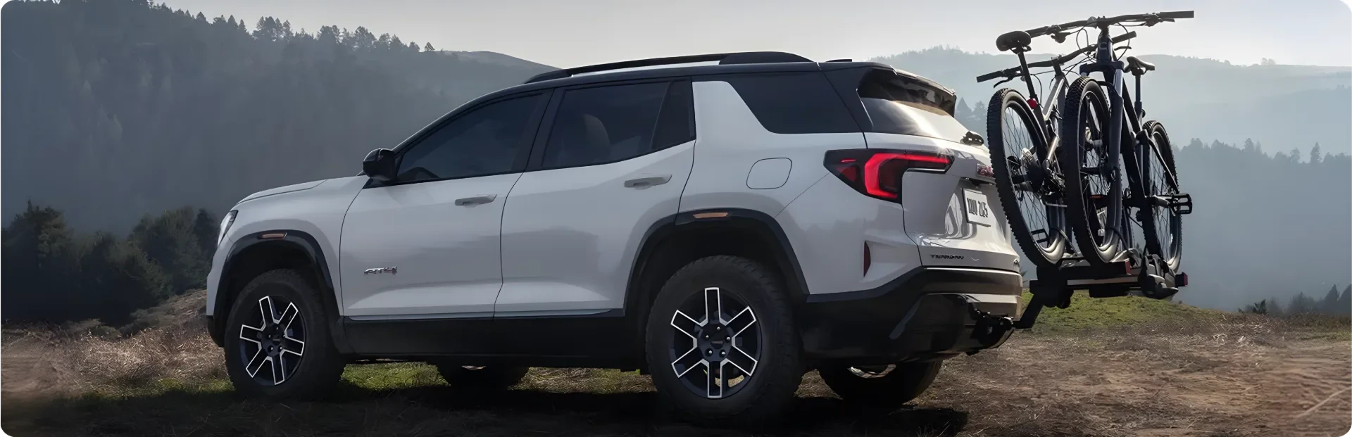 White 2027 GMC Terrain parked on a ridge at dusk with two mountain bikes on a hitch-mounted rack