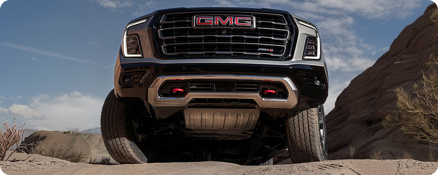 Low-angle front view of a GMC Yukon AT4 Ultimate cresting a sandy rise, skid plate and red recovery hooks visible