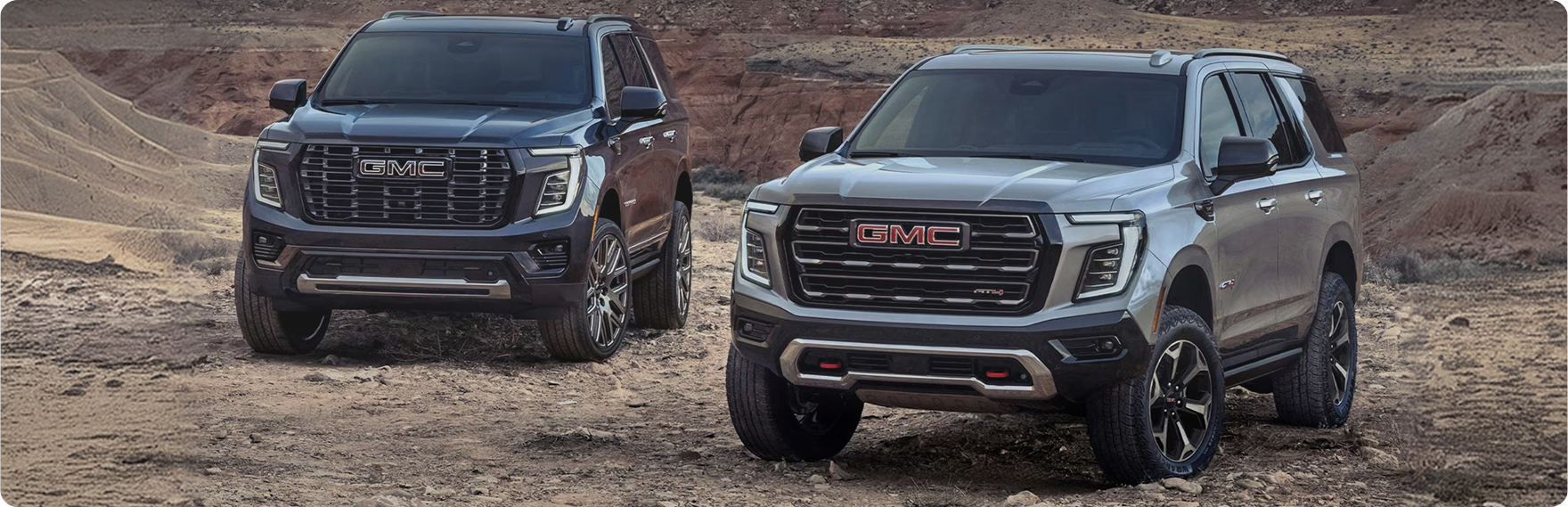 Two GMC Yukon SUVs, a dark Denali and a silver AT4, parked side by side in a desert canyon