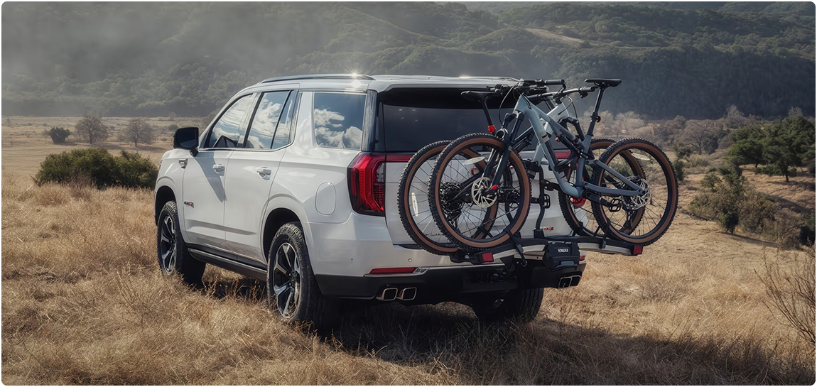White GMC Yukon AT4 carrying two mountain bikes on a hitch rack in a grassy field