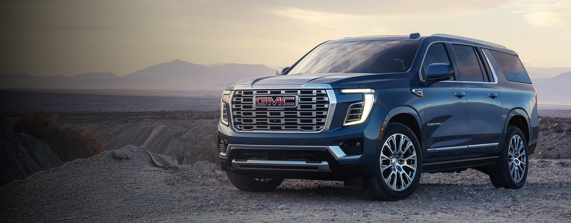 Blue 2027 GMC Yukon Denali parked on a desert overlook at dusk, seen from the front three-quarter angle