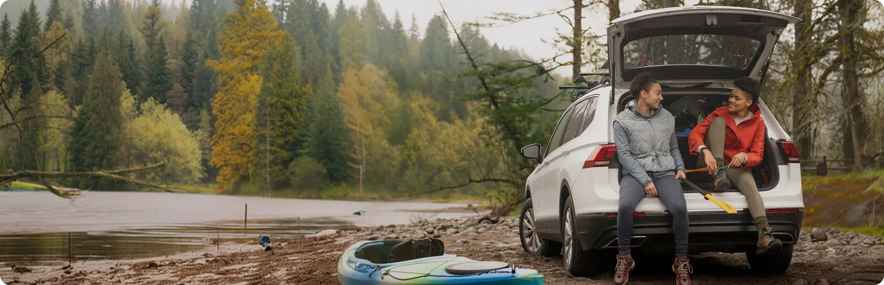 A couple sitting in the open tailgate of a white SUV beside a lake with kayaks on the shore