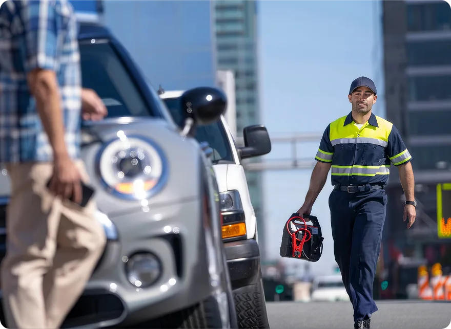 A roadside assistance technician in a hi-vis vest walking toward a stopped vehicle with a jump pack