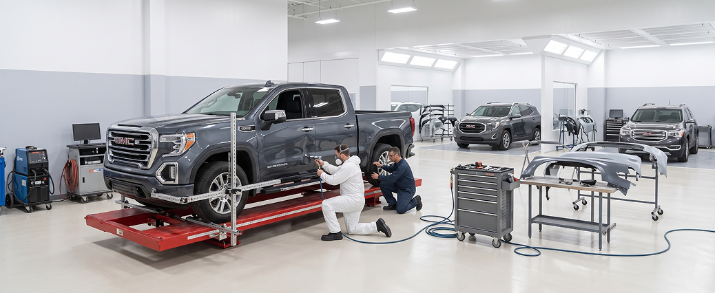 A grey GMC Sierra on a red frame-straightening rack as two technicians work on it, with replacement body panels on stands nearby