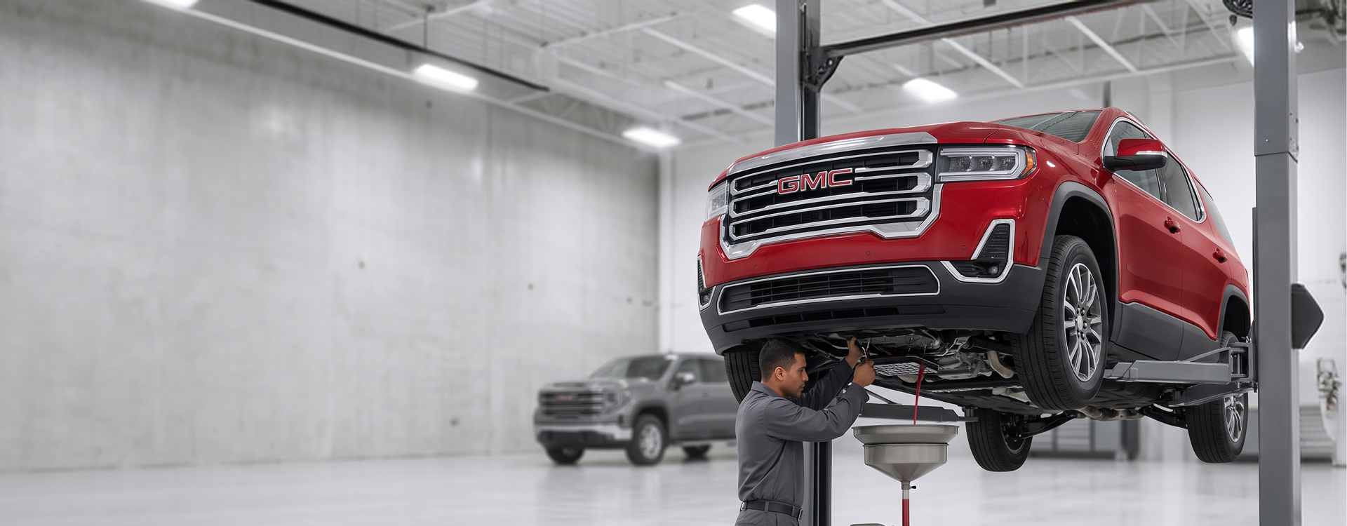 A technician in grey coveralls works beneath a red GMC Acadia raised on a lift as fluid drains into a funnel-topped drain, with a grey GMC Sierra parked behind