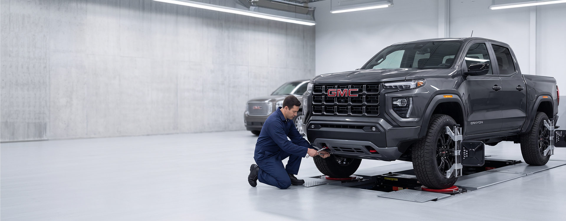 A technician in navy coveralls kneels with a wrench beside a dark grey GMC Canyon on an alignment rack, with wheel clamps and targets fitted to its wheels